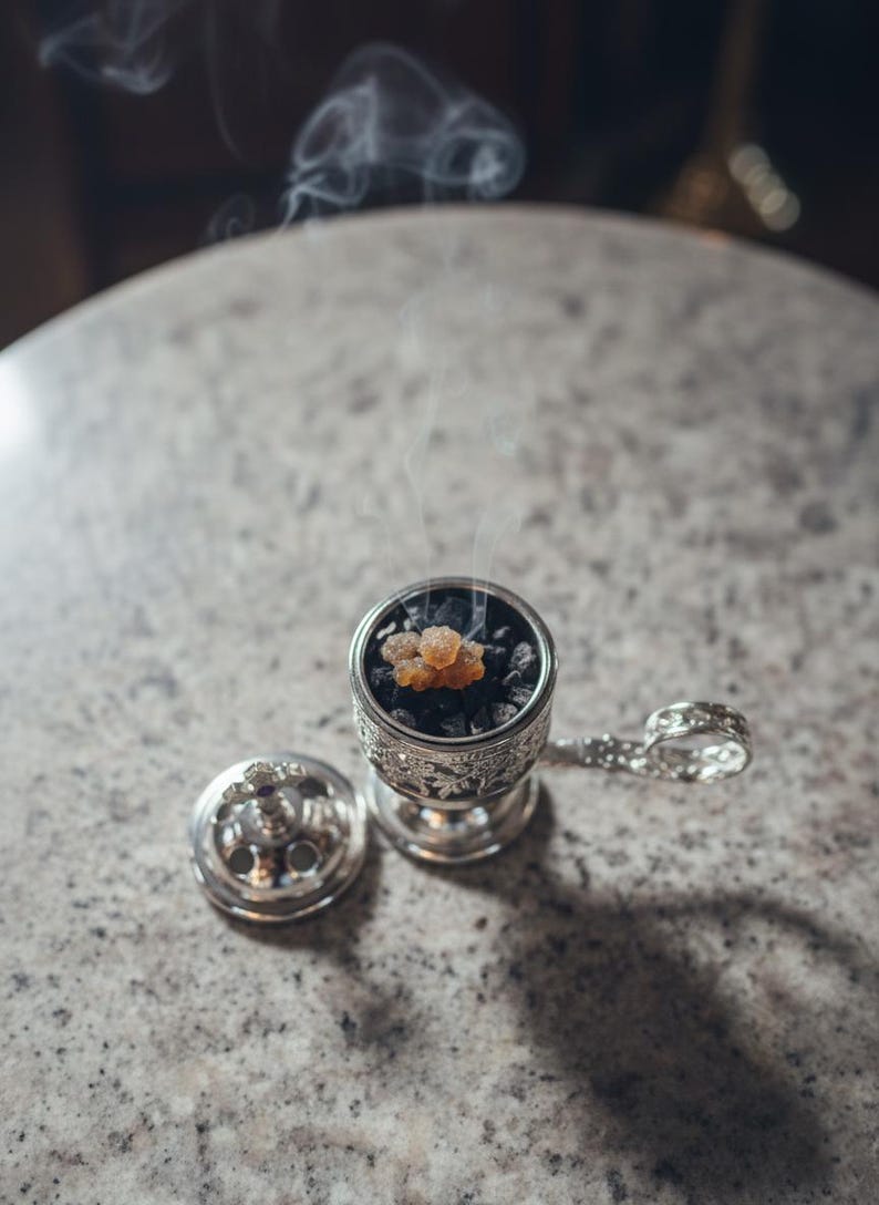 May include: A detailed silver incense burner with a lid and handle, containing dark charcoal and amber resin. Smoke gently rises from the burner, indicating it is lit. The burner is placed on a speckled, light-coloured surface.