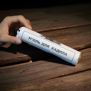 May include: A white paper-wrapped product with black text in Russian, held by a hand. The product is cylindrical and rests on a dark wooden surface. The text on the paper is partially visible.