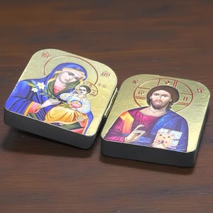 以下が含まれることがあります： ヒンジ付きの2つの金色の宗教アイコン。1つは聖母マリアと子供、もう1つはイエス・キリストを描いています。アイコンは黒い縁取りのある長方形です。アートワークは鮮やかな色と複雑なディテールが特徴です。