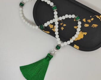 Puede incluir: Un rosario de cuentas blancas con detalles de malaquita verde y una borla verde. El rosario se exhibe en una bandeja negra con detalles de pan de oro. Las cuentas son redondas y están espaciadas uniformemente.