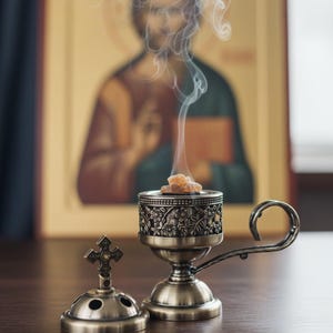 May include: An antique brass incense burner with a floral design, open, with a small cross-shaped lid next to it. Incense is burning, with smoke rising. A religious icon is in the background.
