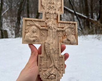 May include: A detailed wooden cross with carved religious figures. The cross shows Jesus on the cross, with figures on either side and detailed carvings at the top and bottom. The cross is held against a snowy, wooded backdrop.