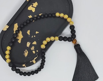 Puede incluir: Un collar de cuentas negras con detalles dorados y una borla negra. El collar se exhibe en una bandeja negra con detalles de pan de oro. Las cuentas tienen aproximadamente 0,8 cm de diámetro.