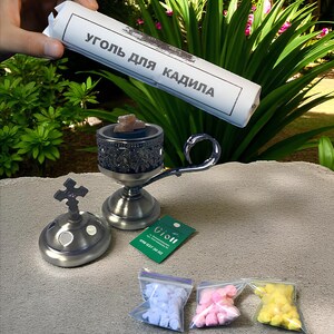May include: A collection of religious items, including a silver incense burner with a cross-shaped top, a small censer, and three small bags of colourful incense. A roll of paper with Cyrillic text is held above the items.