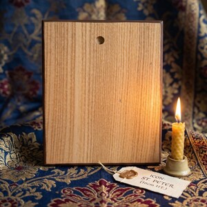May include: Wooden icon of St. Peter, with a light brown wood grain. The icon has a dark brown border and a small hole at the top. A lit beeswax candle sits to the right. A tag reads "ICON: ST. PETER (16cm HT.)".