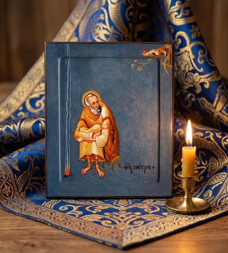 May include: A framed religious icon depicting a saint in orange and yellow robes, seated on a small stool. The icon is set against a blue background with a rooster illustration. A lit candle in a brass holder is in the foreground.