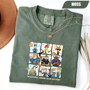 May include: Moss green t-shirt featuring a colorful graphic of characters from Zootopia. The shirt has the text "IN ZOOTOPIA, ANYONE CAN BE ANYTHING". A silver necklace with a pendant is draped on the shirt.