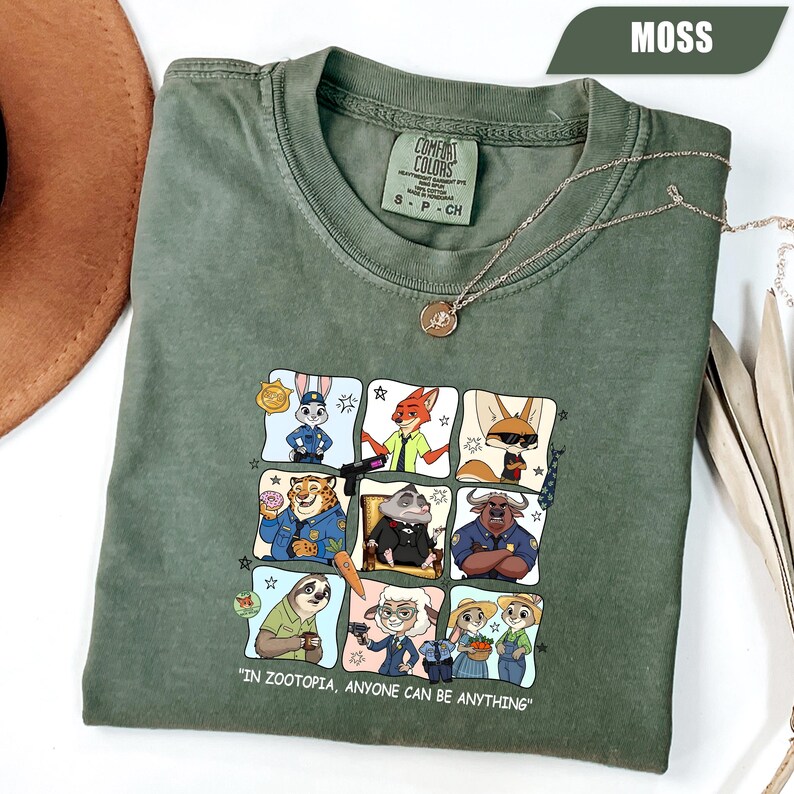 May include: Moss green t-shirt featuring a colorful graphic of characters from Zootopia. The shirt has the text "IN ZOOTOPIA, ANYONE CAN BE ANYTHING". A silver necklace with a pendant is draped on the shirt.