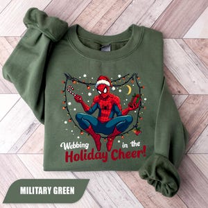 May include: Military green sweatshirt featuring a graphic of Spider-Man in a Santa hat, holding a candy cane and a gift. The text "Webbing in the Holiday Cheer!" is printed below the graphic. The sweatshirt is made of a soft material.