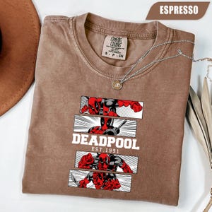 以下が含まれることがあります： エスプレッソ色のTシャツ。デッドプールのグラフィックが特徴です。デザインには、赤と黒のデッドプール、白い「DEADPOOL」、そして「EST.1991」の文字が描かれたコミックスタイルのパネルが含まれています。ペンダント付きのシルバーネックレスがシャツの上にかけられています。