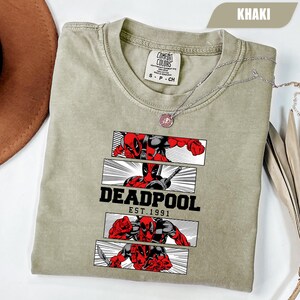 以下が含まれることがあります： カーキ色のTシャツ。デッドプールのコミック風グラフィックが特徴です。デザインには、アクションポーズのデッドプールがパネルに描かれ、「DEADPOOL」の文字と「EST.1991」の文字が配置されています。シルバーのネックレスがTシャツの上にかけられています。