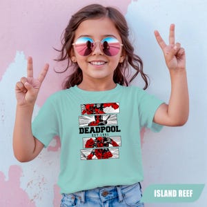 以下が含まれることがあります： 「DEADPOOL EST 1991」の文字とデッドプールのグラフィックが特徴のライトブルーのTシャツ。デザインには、赤と黒のコミックスタイルのイラストが含まれています。Tシャツはサングラスとブルージーンズを着用した人が着ています。