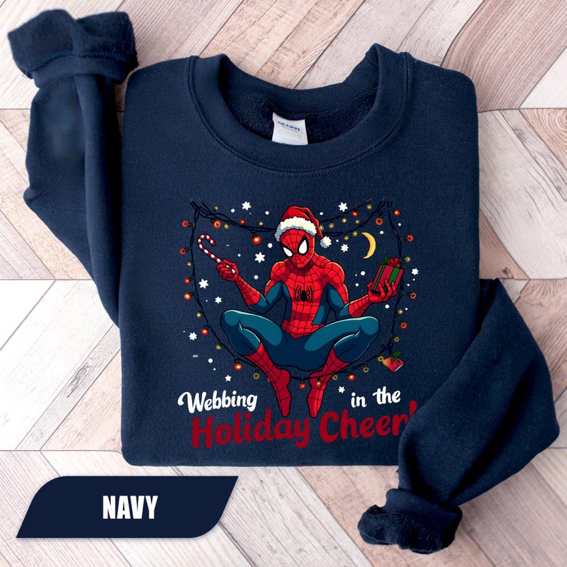 May include: Navy blue sweatshirt featuring a graphic of Spider-Man in a Santa hat, holding a candy cane and a gift. The text "Webbing in the Holiday Cheer" is printed below the graphic. The sweatshirt is made of a soft material.