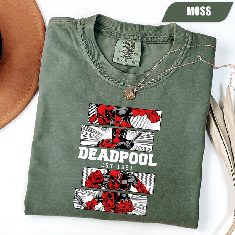 以下が含まれることがあります： モスグリーンのTシャツ。デッドプールのグラフィックが特徴です。赤と黒のデッドプールが描かれたコミック風のパネルがデザインされています。「DEADPOOL EST.1991」の文字が下にプリントされています。金のネックレスがTシャツの上にかけられています。