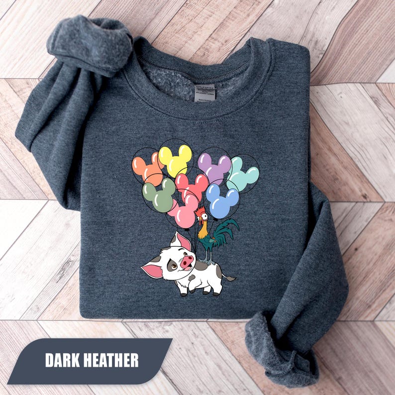 May include: Dark heather sweatshirt featuring a cartoon pig holding colorful Mickey Mouse-shaped balloons, with a rooster perched on its back. The text "DARK HEATHER" is displayed at the bottom. The sweatshirt is folded on a wooden surface.