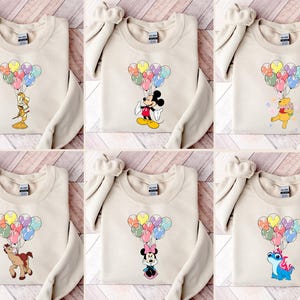 May include: Beige sweatshirts featuring various cartoon characters holding colorful balloon bouquets. Characters include Mickey Mouse, Minnie Mouse, Winnie the Pooh, and others. The sweatshirts are displayed on a wooden surface.