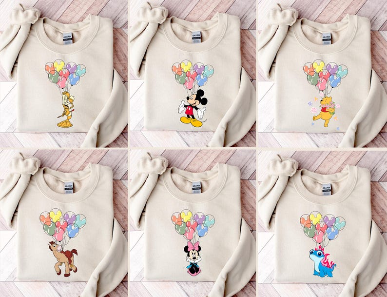 May include: Beige sweatshirts featuring various cartoon characters holding colorful balloon bouquets. Characters include Mickey Mouse, Minnie Mouse, Winnie the Pooh, and others. The sweatshirts are displayed on a wooden surface.