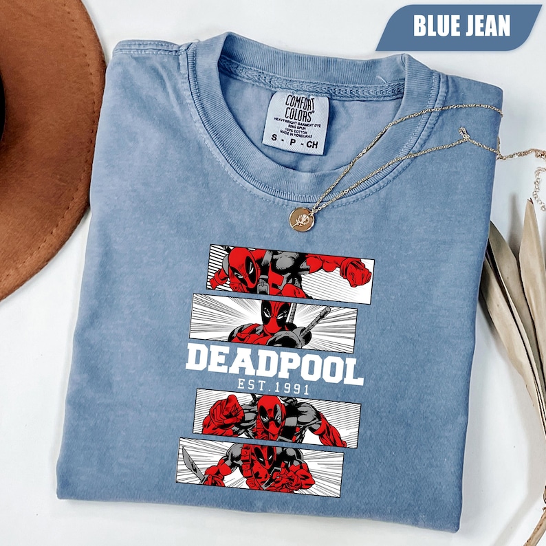 以下が含まれることがあります： 赤と黒のデッドプールのグラフィックが特徴の、ブルーデニムカラーのTシャツ。シャツには白いブロック体で「DEADPOOL」の文字があり、その下に「EST. 1991」と書かれています。金のネックレスがシャツの上にかけられています。