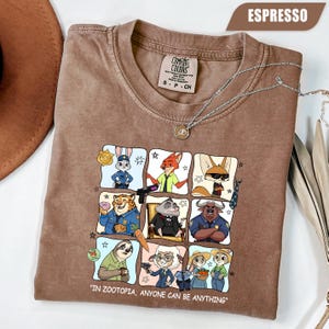 May include: Espresso-colored t-shirt featuring a graphic of Zootopia characters in a grid. The shirt includes the text "IN ZOOTOPIA, ANYONE CAN BE ANYTHING." A silver necklace with a pendant is draped across the shirt.
