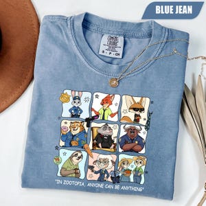 May include: Blue jean colored t-shirt featuring a colorful graphic of characters from Zootopia. The shirt has the text "IN ZOOTOPIA, ANYONE CAN BE ANYTHING". A gold necklace is draped across the shirt.