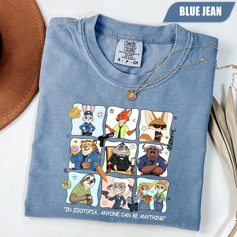 May include: Blue jean colored t-shirt featuring a colorful graphic of characters from Zootopia. The shirt has the text "IN ZOOTOPIA, ANYONE CAN BE ANYTHING". A gold necklace is draped across the shirt.