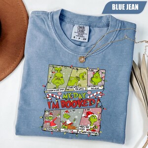 Peut inclure: T-shirt bleu jean Comfort Colors avec un motif de bande dessinée du Grinch. Le texte sur le t-shirt indique "My Day I'm Booked" avec des activités sur le thème du Grinch. Un collier doré est drapé sur le t-shirt.