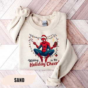 May include: Sand-colored sweatshirt featuring a graphic of Spider-Man in a Santa hat, holding a candy cane and a gift. The design includes string lights and the text "Webbing in the Holiday Cheer".