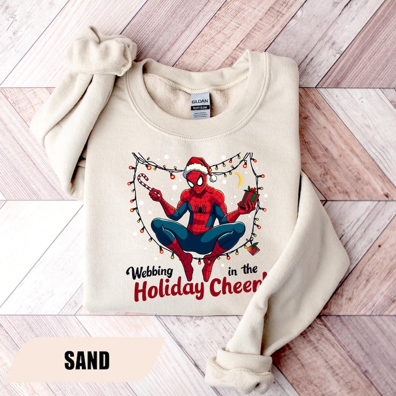 May include: Sand-colored sweatshirt featuring a graphic of Spider-Man in a Santa hat, holding a candy cane and a gift. The design includes string lights and the text "Webbing in the Holiday Cheer".