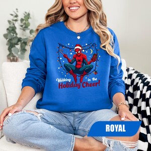 May include: Royal blue sweatshirt featuring a graphic of Spider-Man in a Santa hat, surrounded by Christmas lights and holding a candy cane. The text "Webbing in the Holiday Cheer!" is printed below the graphic.