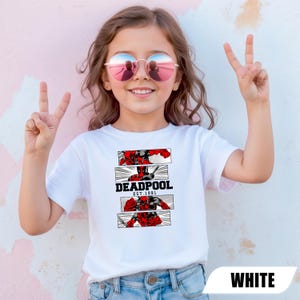 以下が含まれることがあります： 赤と黒のDeadpoolグラフィックと「DEADPOOL EST. 1991」の文字が入った白いTシャツ。Tシャツを着ている人はサングラスをかけ、ピースサインをしています。
