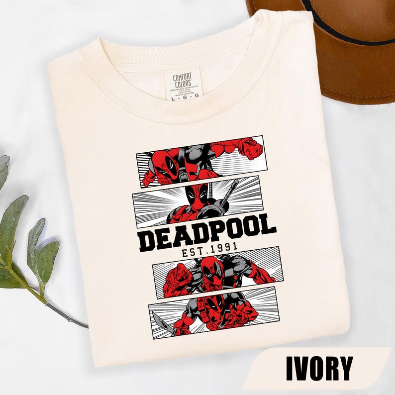 以下が含まれることがあります： アイボリー色のTシャツ。赤と黒のデッドプールのコミック風グラフィックが特徴で、黒で「DEADPOOL EST. 1991」と書かれています。シャツは折りたたまれ、ニュートラルな背景に表示されています。