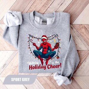 May include: Sport grey sweatshirt featuring a Christmas-themed Spider-Man graphic. The superhero is wearing a Santa hat and holding a candy cane and a gift. The text reads "Webbing in the Holiday Cheer!"