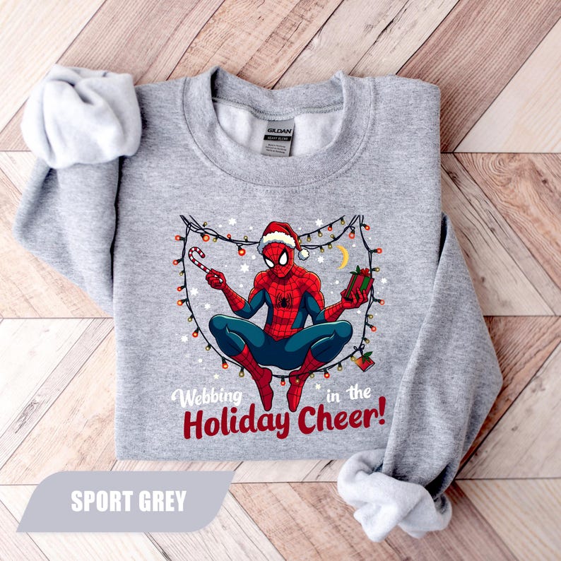 May include: Sport grey sweatshirt featuring a Christmas-themed Spider-Man graphic. The superhero is wearing a Santa hat and holding a candy cane and a gift. The text reads "Webbing in the Holiday Cheer!"