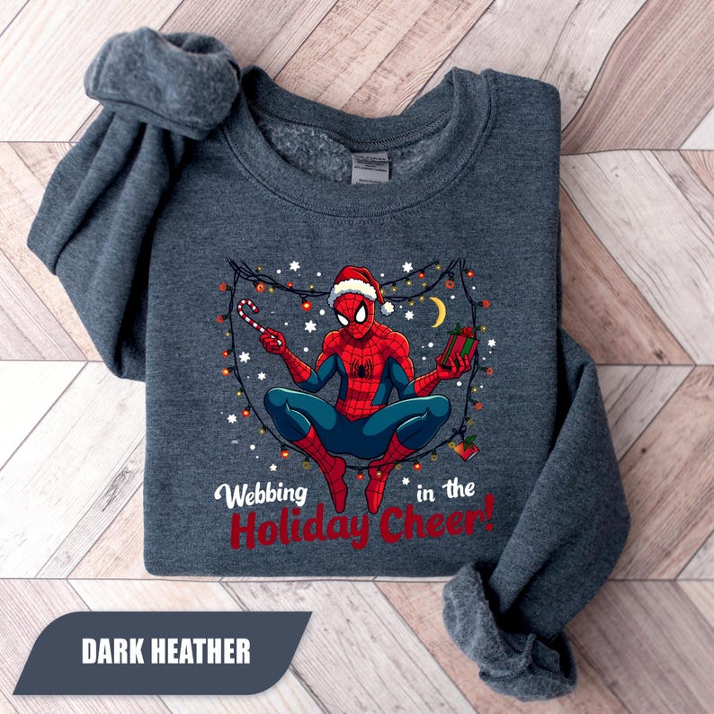 May include: Dark heather gray sweatshirt featuring a graphic of Spider-Man in a Santa hat, holding a candy cane and a gift. The text "Webbing in the Holiday Cheer!" is printed below the graphic. The sweatshirt is folded.