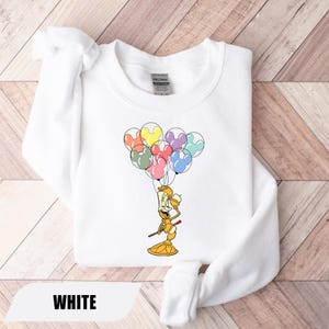 May include: White sweatshirt featuring a colorful cartoon illustration of a candlestick character holding balloons shaped like Mickey Mouse ears. The word "WHITE" is printed at the bottom. The sweatshirt is made of a soft material.