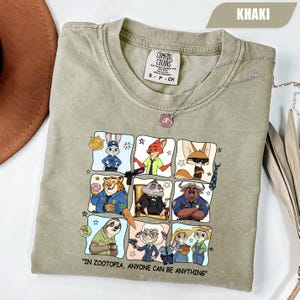 May include: Khaki-colored t-shirt featuring a grid of Zootopia characters. The shirt includes the text "IN ZOOTOPIA, ANYONE CAN BE ANYTHING." A silver necklace with a pendant is draped across the shirt. The shirt is made of Comfort Colors material.