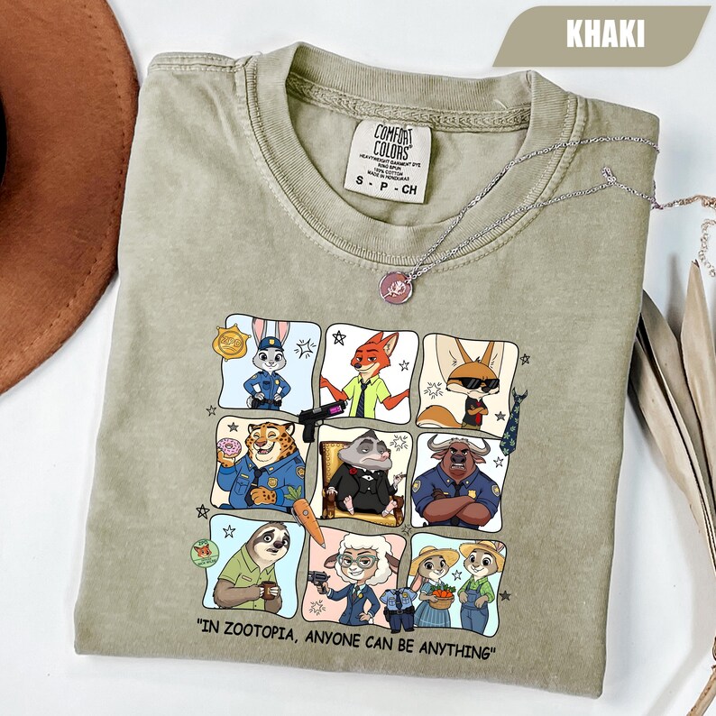 May include: Khaki-colored t-shirt featuring a grid of Zootopia characters. The shirt includes the text "IN ZOOTOPIA, ANYONE CAN BE ANYTHING." A silver necklace with a pendant is draped across the shirt. The shirt is made of Comfort Colors material.