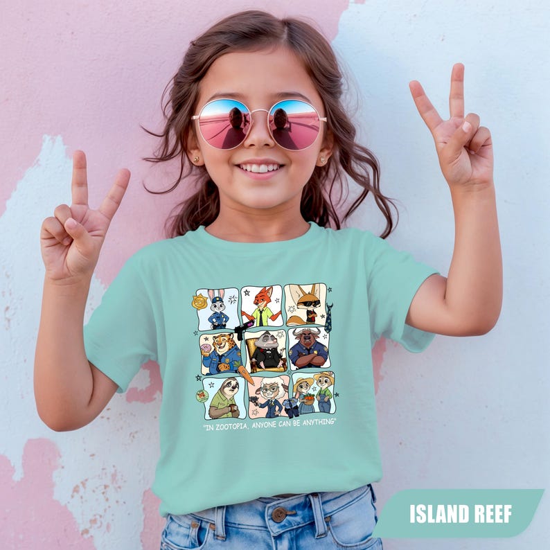 May include: A light blue t-shirt featuring a grid of cartoon characters from Zootopia. The shirt has the text "IN ZOOTOPIA, ANYONE CAN BE ANYTHING". The girl is wearing round sunglasses and making a peace sign.