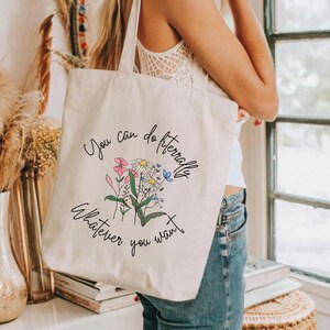 May include: A cream tote bag featuring the phrase "You can do literally whatever you want" and a floral illustration. The flowers are pink, white, and blue, with green stems. The bag has long straps for carrying.