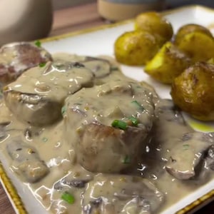 Könnte beinhalten: Nahaufnahme eines Tellers mit cremiger Champignonsauce über Fleischmedaillons, garniert mit grünen Kräutern. Dazu werden Bratkartoffeln serviert. Das Gericht wird auf einem rechteckigen Teller mit Goldrand serviert.