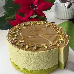 Può includere: Una torta rotonda al pistacchio con una copertura al caramello e pistacchi tritati. Una fetta è stata tagliata dalla torta, rivelando un interno a strati. La torta è su un piatto bianco, con fiori rossi sullo sfondo.