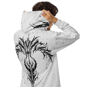 Pode incluir: Moletom branco com um gráfico preto estilizado nas costas. O design é um padrão simétrico intrincado. O moletom tem mangas compridas e um capuz. O tecido tem uma aparência desgastada e salpicada.