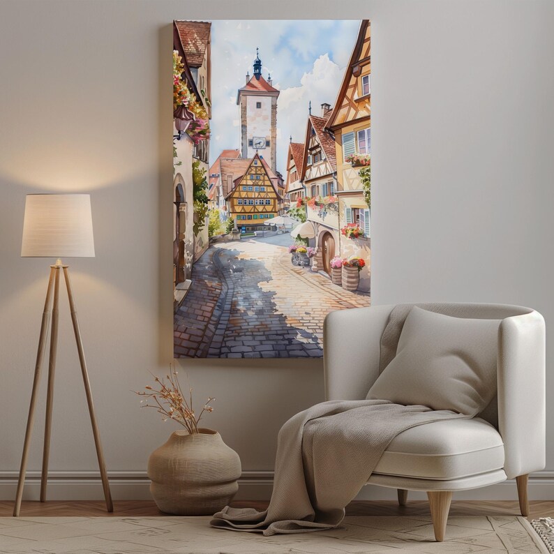 May include: Watercolor painting of a European town street scene. The artwork depicts cobblestone streets, colorful buildings, and a tall clock tower. The painting is displayed on a wall above a white armchair.
