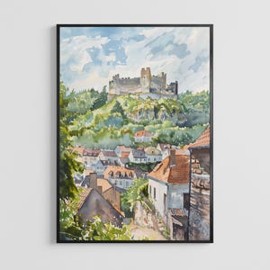 May include: A framed watercolour painting depicting a stone castle perched atop a lush green hill overlooking a town with houses featuring orange and grey roofs. The sky is filled with soft blue and white clouds above the dense forest landscape.