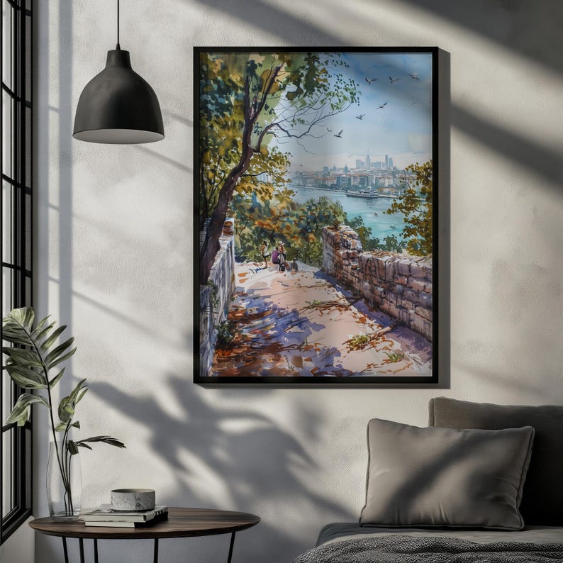 Könnte beinhalten: Ein gerahmtes Aquarellgemälde zeigt eine malerische Aussicht auf eine Stadt am Fluss. Das Kunstwerk zeigt einen Steinweg, üppige Bäume und eine entfernte Stadtlandschaft unter blauem Himmel. Das Gemälde befindet sich in einem schwarzen Rahmen.