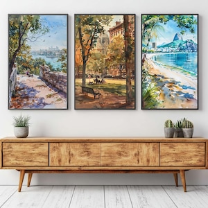 May include: Three framed watercolor paintings depicting scenic landscapes. One shows a coastal view with a city skyline, another a park scene with a bench, and the third a beach with a mountain backdrop. The frames are black, and the artwork is displayed above a wooden cabinet.