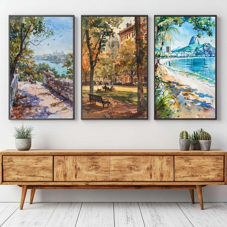 May include: Three framed watercolor paintings depicting scenic landscapes. One shows a coastal view with a city skyline, another a park scene with a bench, and the third a beach with a mountain backdrop. The frames are black, and the artwork is displayed above a wooden cabinet.