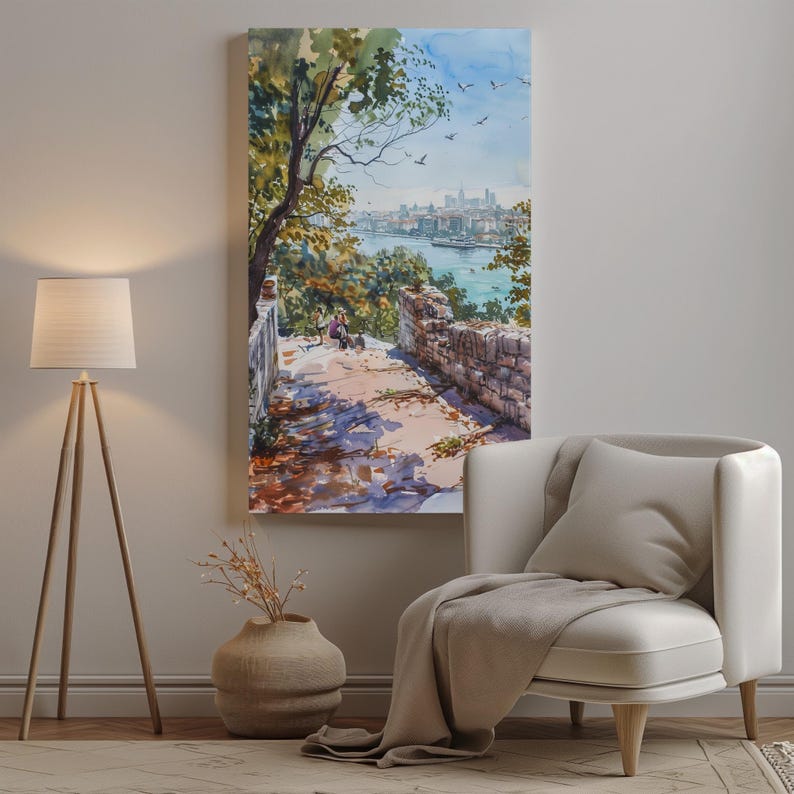 May include: A watercolor painting depicts a scenic overlook with a stone wall, path, and trees. The artwork showcases a river and city skyline in the distance, with a blue sky and birds. The painting is displayed on a wall above a white armchair.