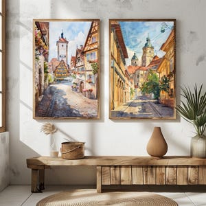 May include: Two framed watercolor prints of European street scenes. The artwork depicts cobblestone streets, colorful buildings, and a church tower. The frames are a warm brown color, and the overall style is reminiscent of travel art.
