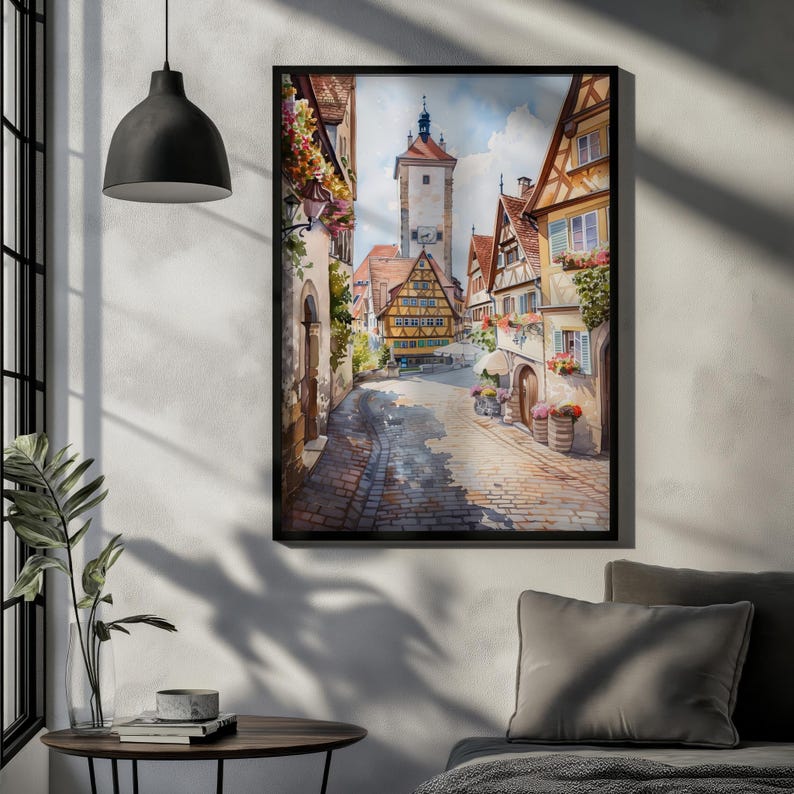 Könnte beinhalten: Gerahmter Aquarelldruck einer europäischen Stadtstraßenszene. Das Kunstwerk zeigt Kopfsteinpflasterstraßen, farbenfrohe Gebäude mit Blumen und einen hohen Turm im Hintergrund. Der Druck wird an einer weißen Wand ausgestellt.